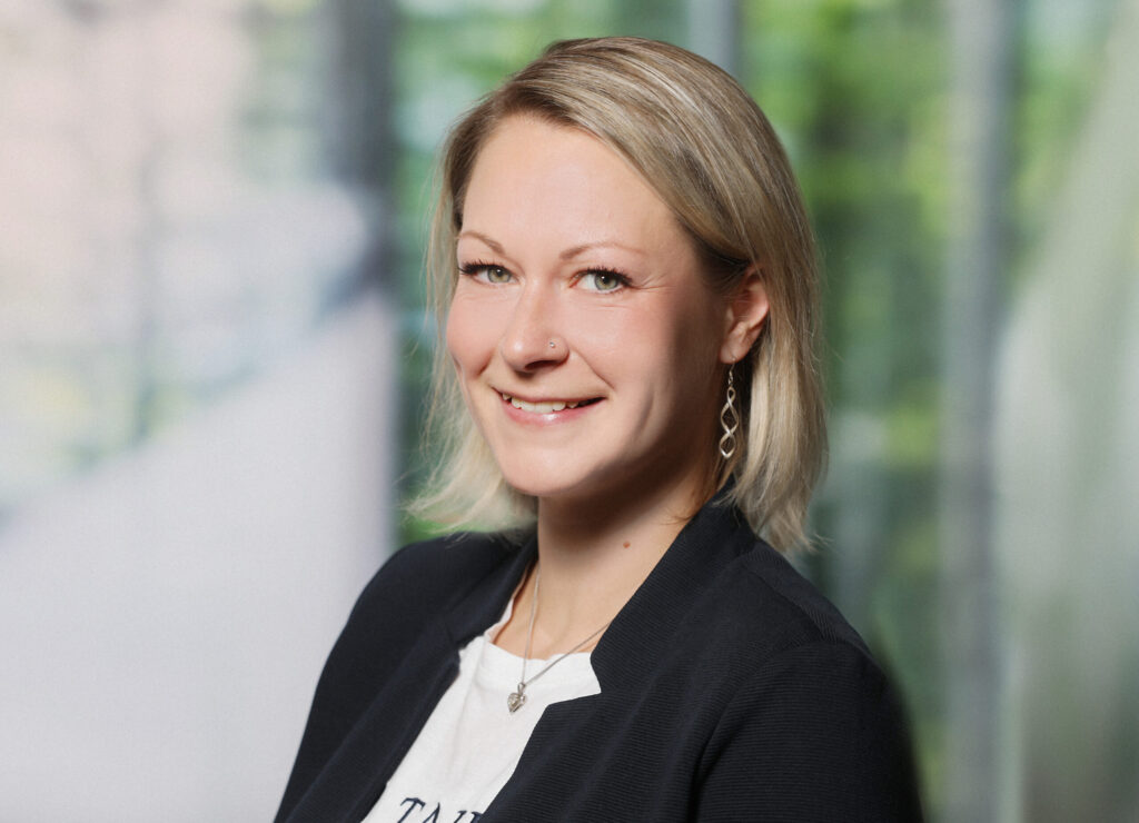 Porträt einer Frau mit hellem Haar in Businesskleidung vor unscharfem, grünem Hintergrund.