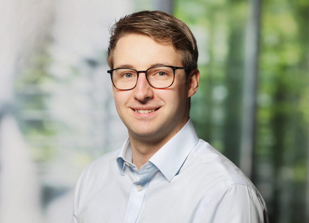 Porträt eines Mannes mit Brille und hellem Hemd vor unscharfem, grünem Hintergrund.